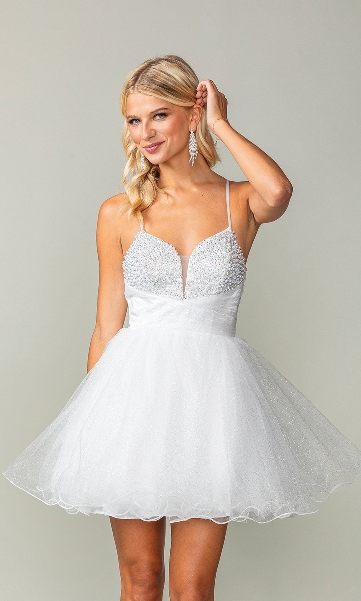 A woman wearing the Lace-Up Short Babydoll Homecoming Dress 3385—white, sparkly, with thin straps and a fitted bodice—stands against a plain background, touching her hair and smiling.